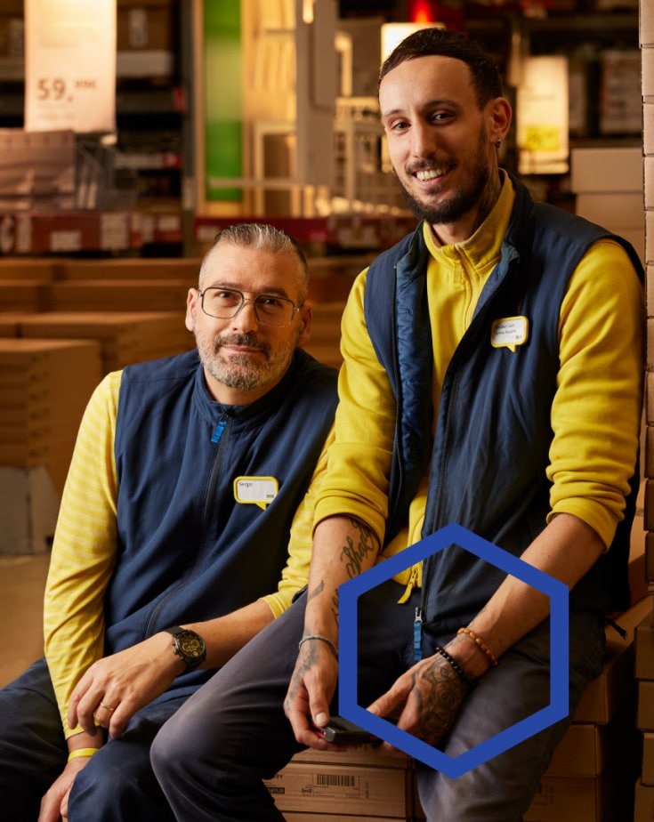 Two IKEA co-workers sitting on flat packs in a stores warehouse.