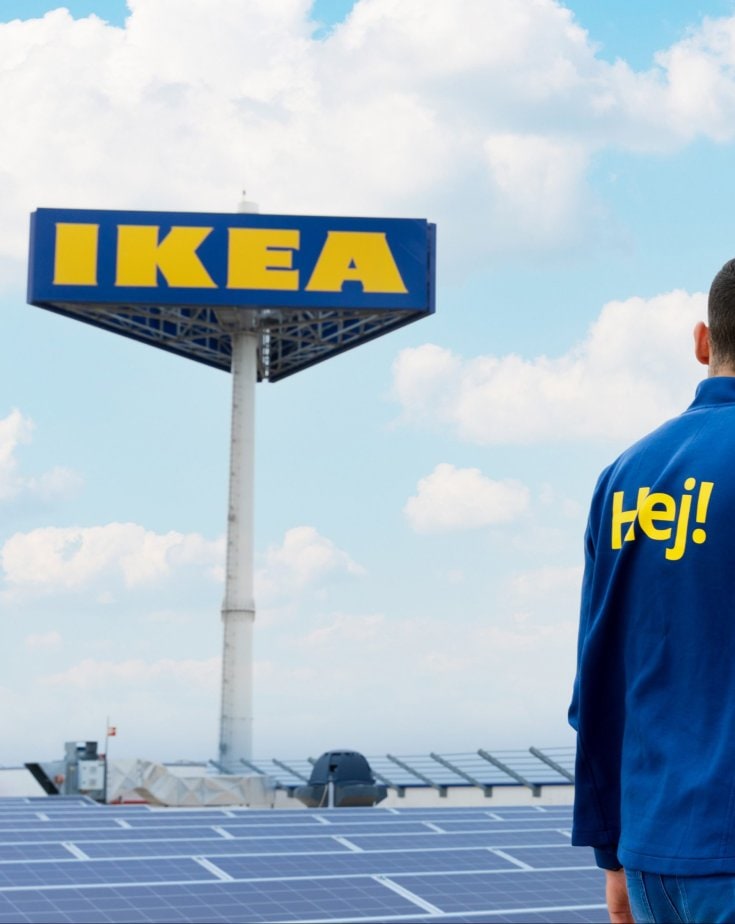 An IKEA employee standing on a roof with solar panels looking up to the store'IKEA sign with a blue, semi-cloudy sky in the background.