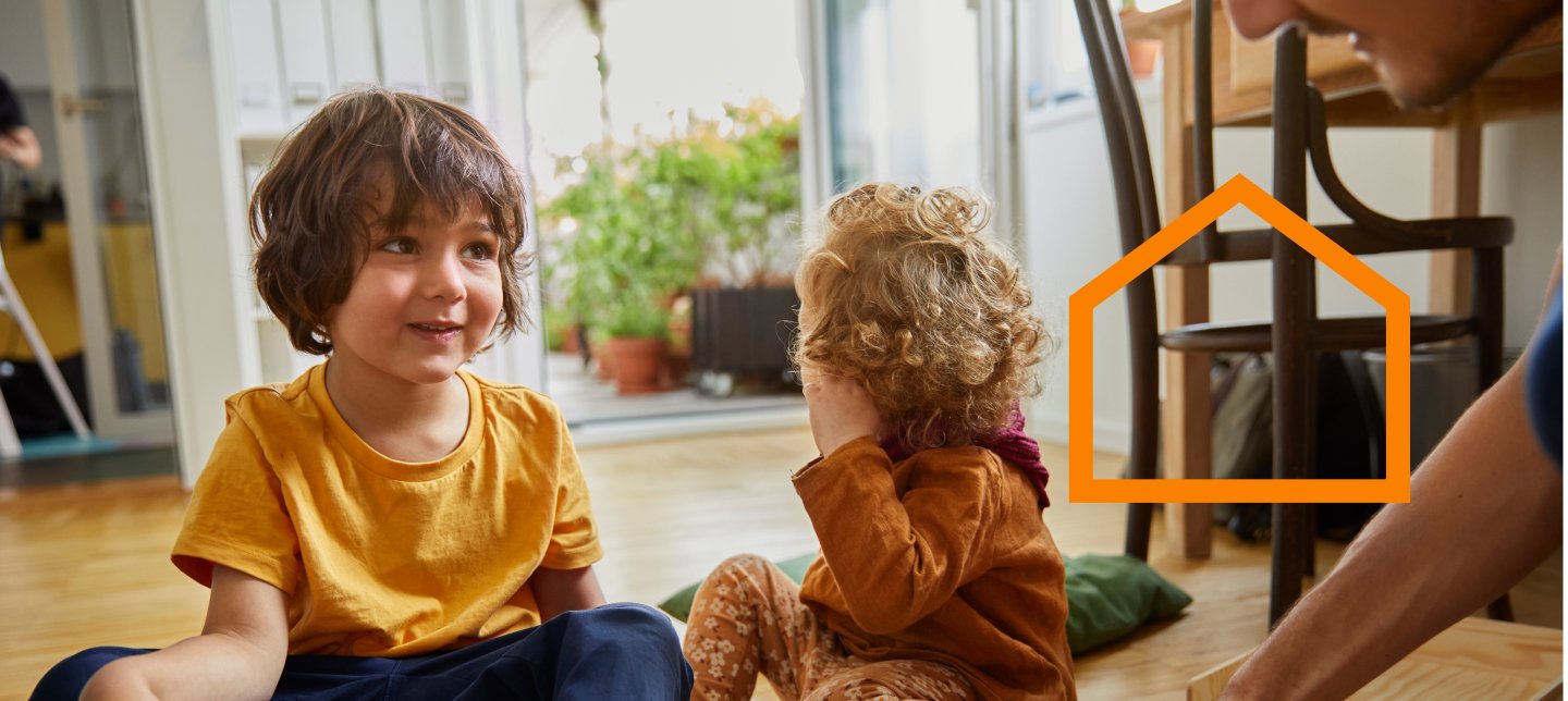 Two small children sits on the livingroom floor while their parent is assembling a piece of IKEA furniture.