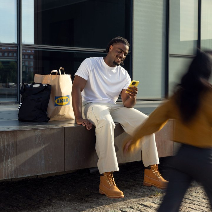 A man sits outside on a ledge looking at his phone smiling with an IKEA paper bag next to him.