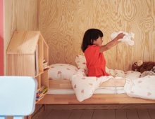 A child wearing a red dress sits on a bed playing with the soft toy SKOGSDUVA while smiling big.