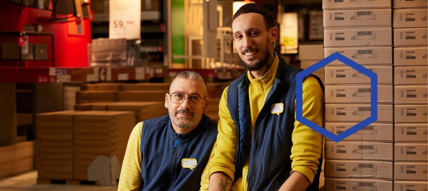 Two IKEA co-workers sitting on flat packs in a stores warehouse.