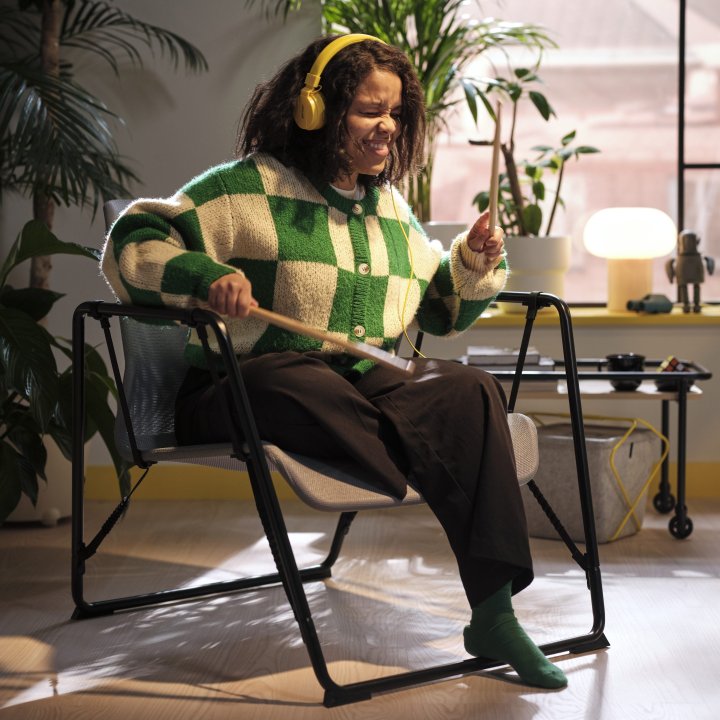 A woman wearing a green and white cardigan plays air drums with headphones on, sitting in a chair from the BRÄNNBOLL collection.