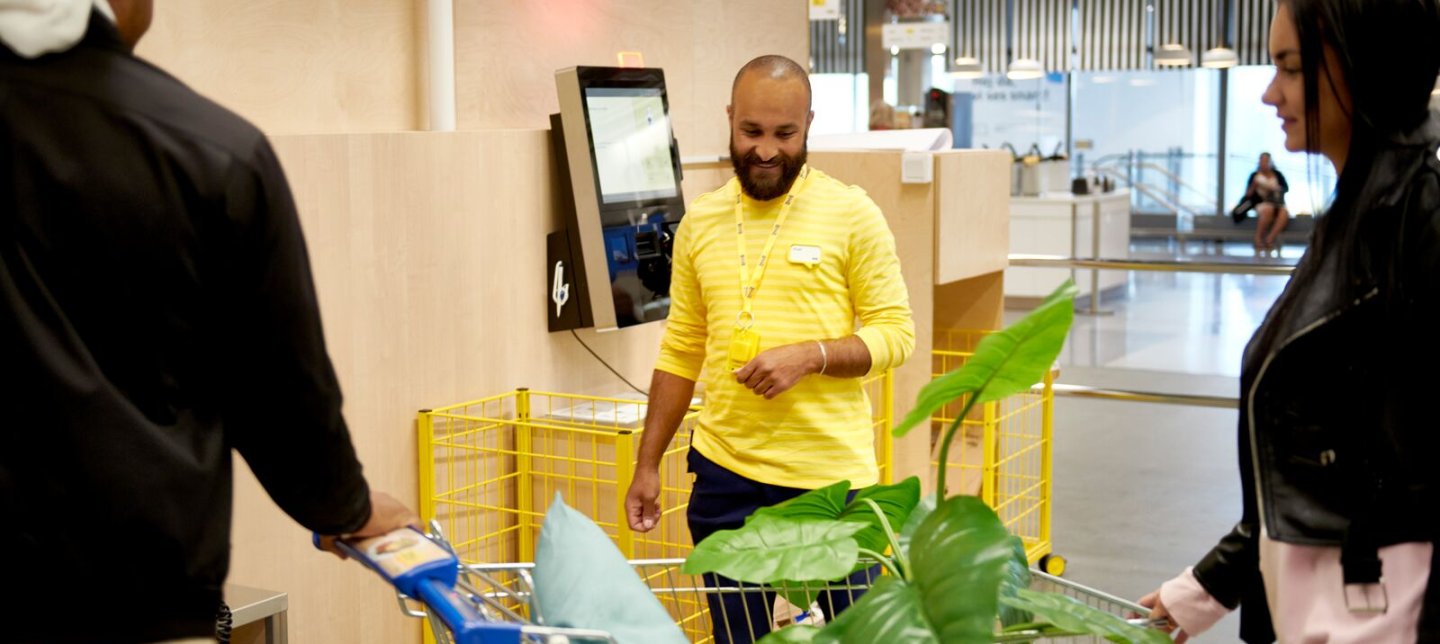 A smiling IKEA employee helping two customers in the self service check-out area.