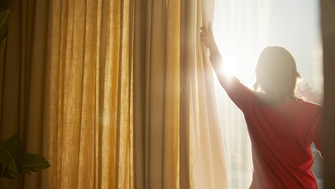 Person pulling curtains aside, allowing the sun to shine in through the windows.