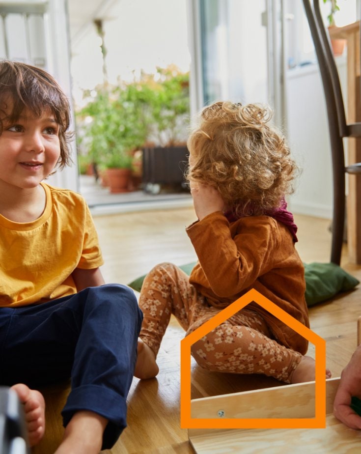 Two small children sits on the livingroom floor while their parent is assembling a piece of IKEA furniture.