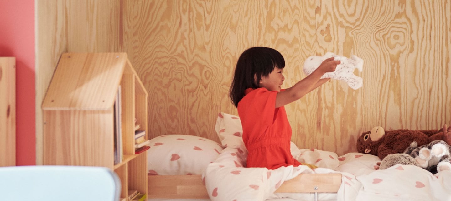 A child wearing a red dress sits on a bed playing with the soft toy SKOGSDUVA while smiling big.