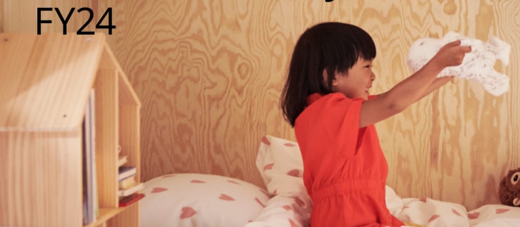 A child wearing a red dress sits on a bed playing with the soft toy SKOGSDUVA while smiling big.