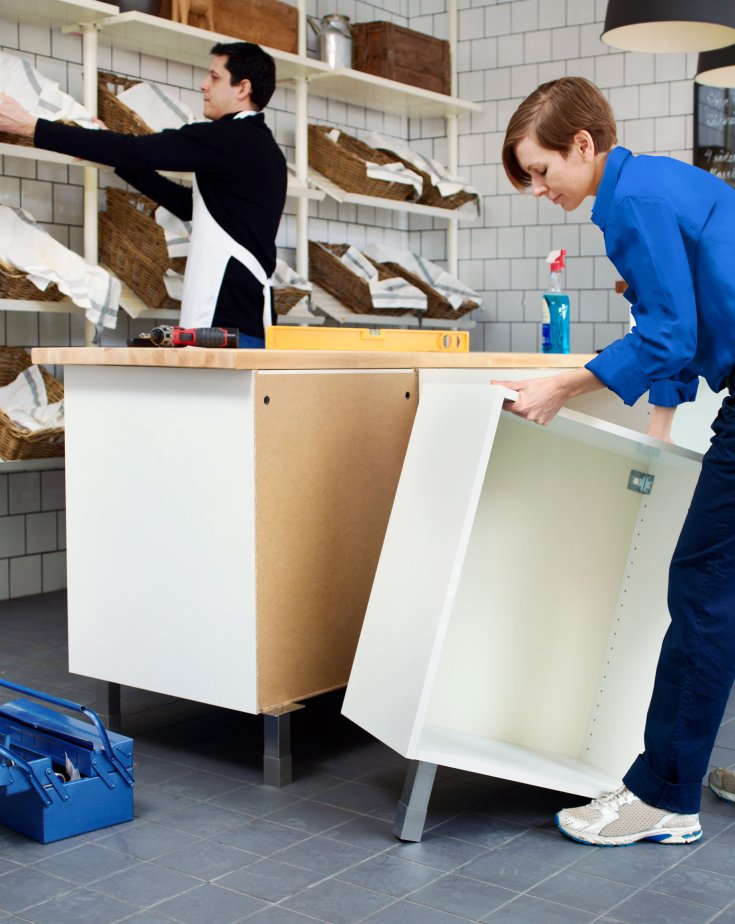 Two IKEA employees putting cabinets together and placing bread baskets on a shelf, decorating a small shop area.